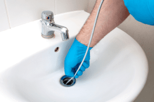 A man's hand wearing blue gloves holding the edge of a plumbing snake which he is threading down the drain